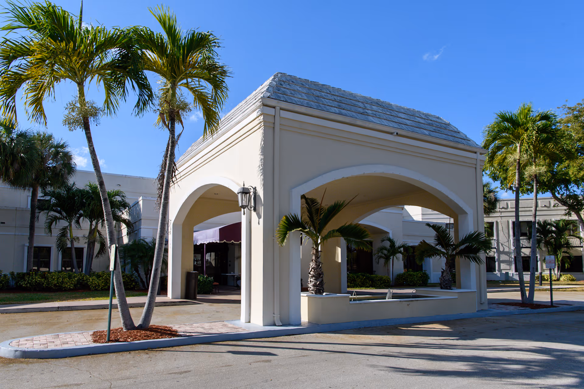 Covered porte-cochère and entrance of a light-colored building framed by palm trees under a clear blue sky.