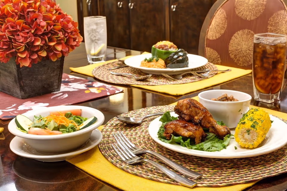 A dining table set with two plates of food, including grilled chicken drumsticks, corn on the cob, a bowl of beans, a stuffed green pepper with rice and beans, and a fresh salad with cucumber and tomato slices. There are two glasses of beverages, one with iced tea and the other with water and lemon. A decorative flower arrangement in a square pot is also on the table.