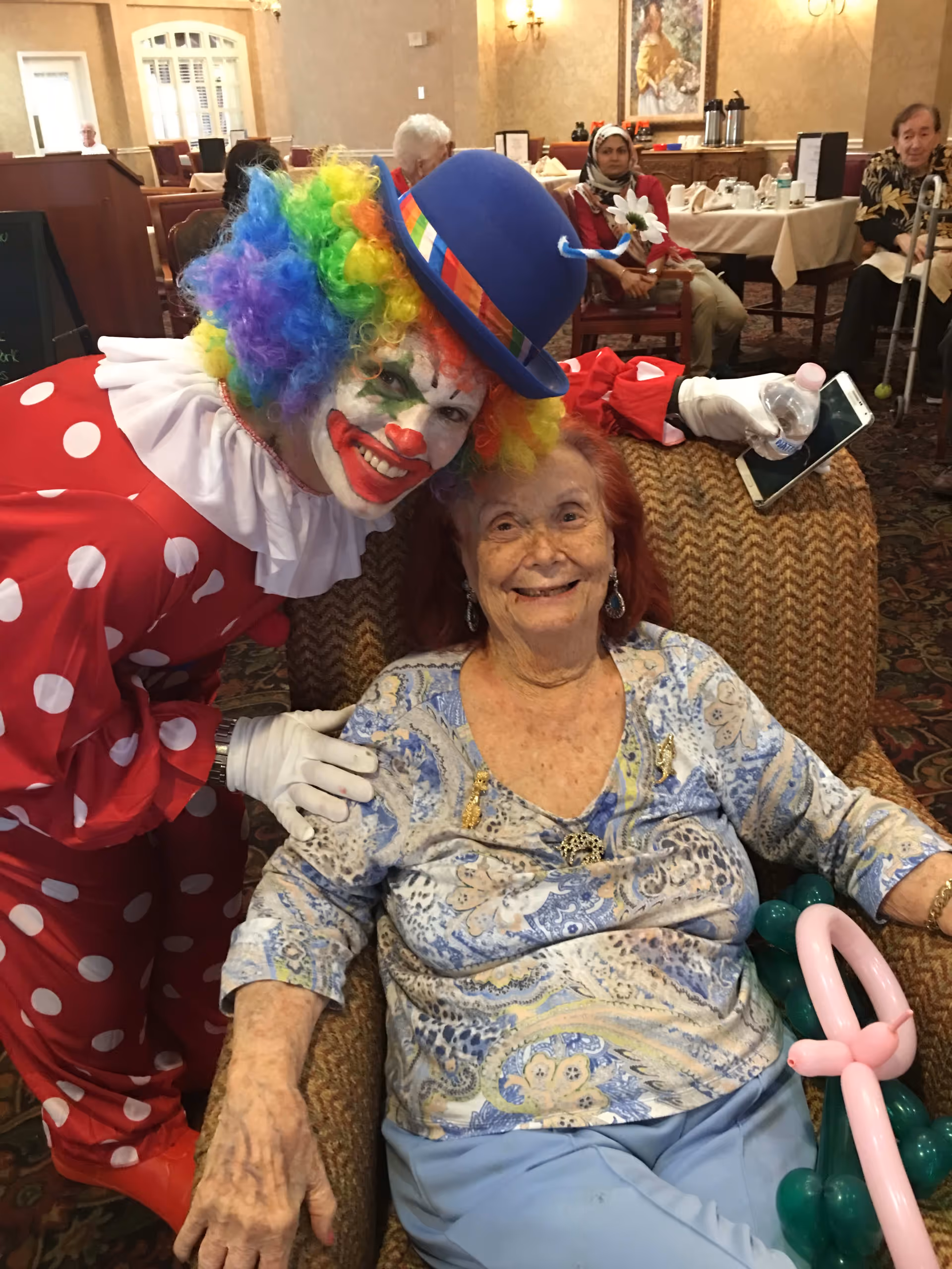 An elderly woman sitting in a patterned armchair smiling, holding a balloon sculpture. Next to her is a person dressed as a clown with a colorful rainbow wig, white face paint, red nose, and a blue hat with a rainbow band, leaning in and smiling. In the background, other elderly people are seated at tables in a dining room setting.