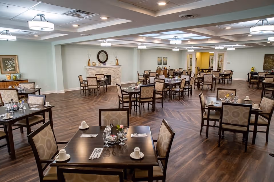 A spacious dining room with multiple dark wood tables and cushioned chairs arranged neatly. Each table is set with cups, glasses, napkins, and silverware. The room features wood-patterned flooring, soft lighting from ceiling fixtures, and a decorative fireplace with a round mirror above it. Walls are painted light green with framed artwork and decorative items on side tables.
