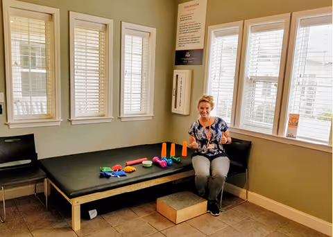 A woman sitting on a black cushioned bench in a well-lit room with several windows. On the bench are colorful small exercise equipment including hand weights and cones. The walls are painted light green and there is a small step stool on the floor in front of the bench.