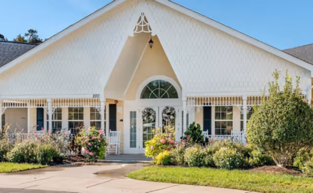 Front exterior view of a single-story building with white siding, a peaked roof, and a covered entrance decorated with floral wreaths on the double doors. The entrance is flanked by windows and surrounded by well-maintained bushes and flowering plants under a clear blue sky.