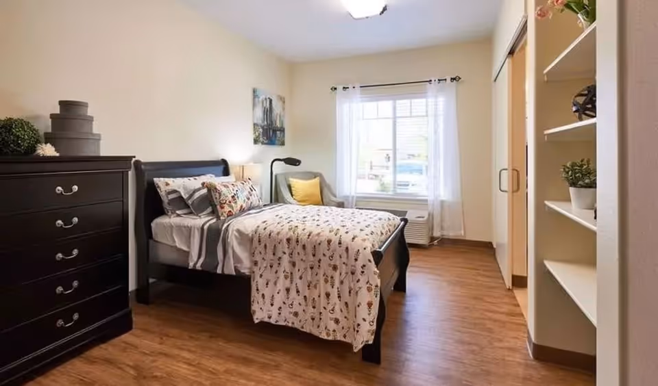 A cozy bedroom with a single bed featuring patterned bedding and multiple pillows. Next to the bed is a black dresser with decorative items on top. A comfortable armchair with a yellow pillow is placed near a window with sheer white curtains. The room has wooden flooring and built-in shelves on the right side.