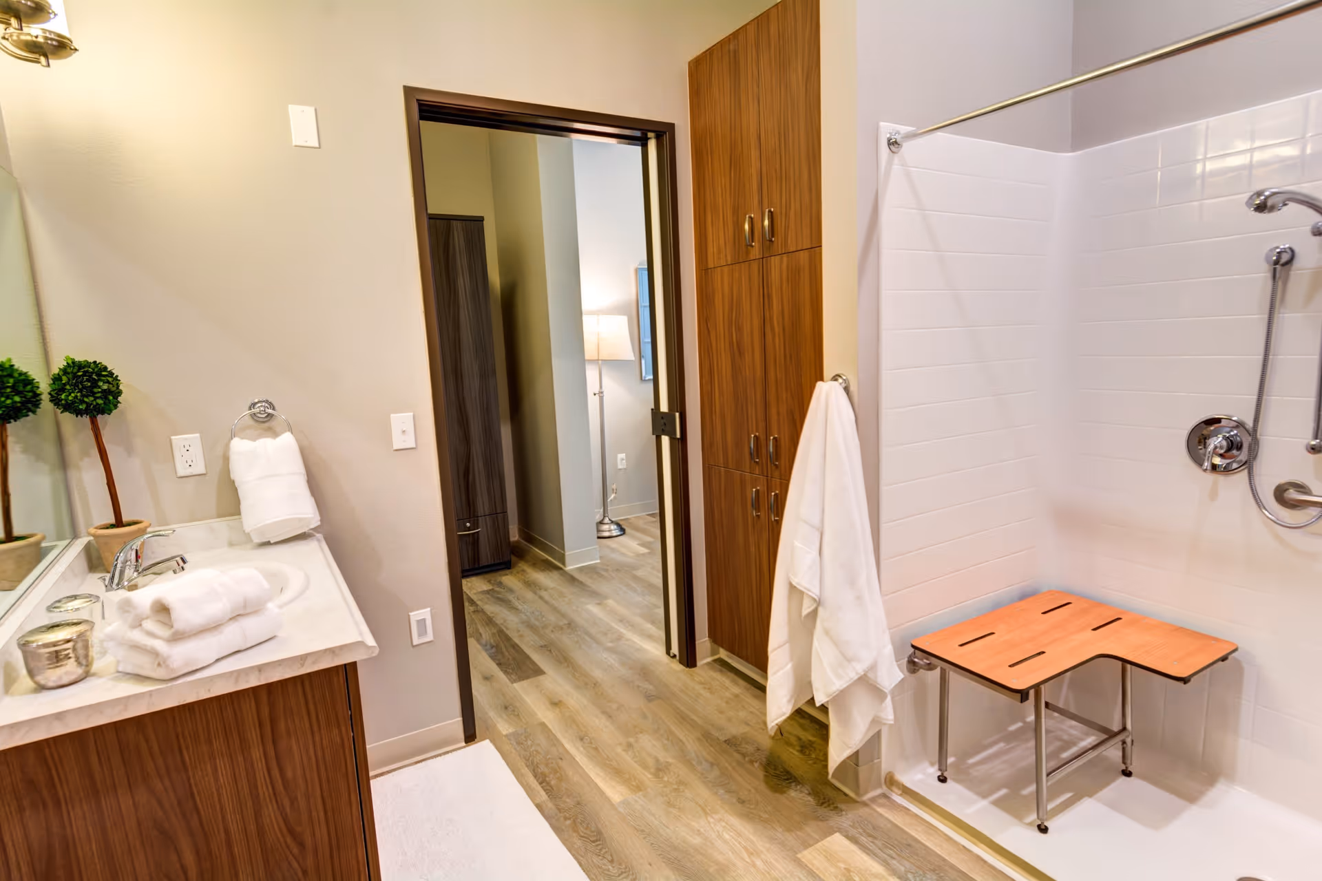 A bathroom with a wooden vanity topped with a white countertop and a sink. Towels and small decorative plants are placed on the countertop. To the right, there is a walk-in shower with white tiled walls and a wooden shower bench. A white towel hangs on a hook near the shower. The bathroom floor is wood-style, and there is a doorway leading to another room with a lamp and furniture visible.