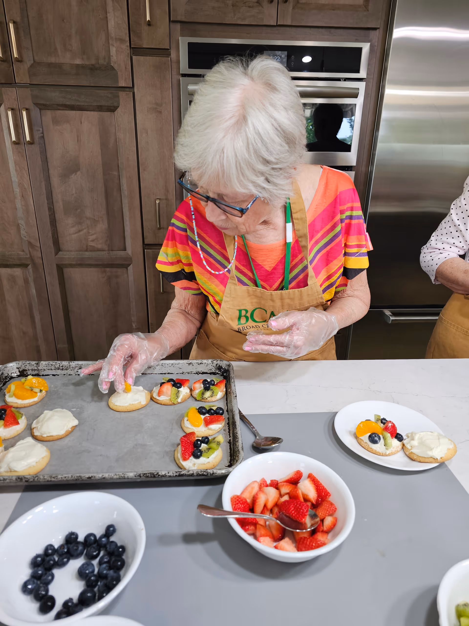 An elderly woman wearing glasses and a colorful striped shirt with a Broad Creek Manor apron is preparing fruit-topped cookies in a kitchen. She is placing fruit on cookies arranged on a baking tray. Bowls of blueberries and sliced strawberries are on the counter in front of her.