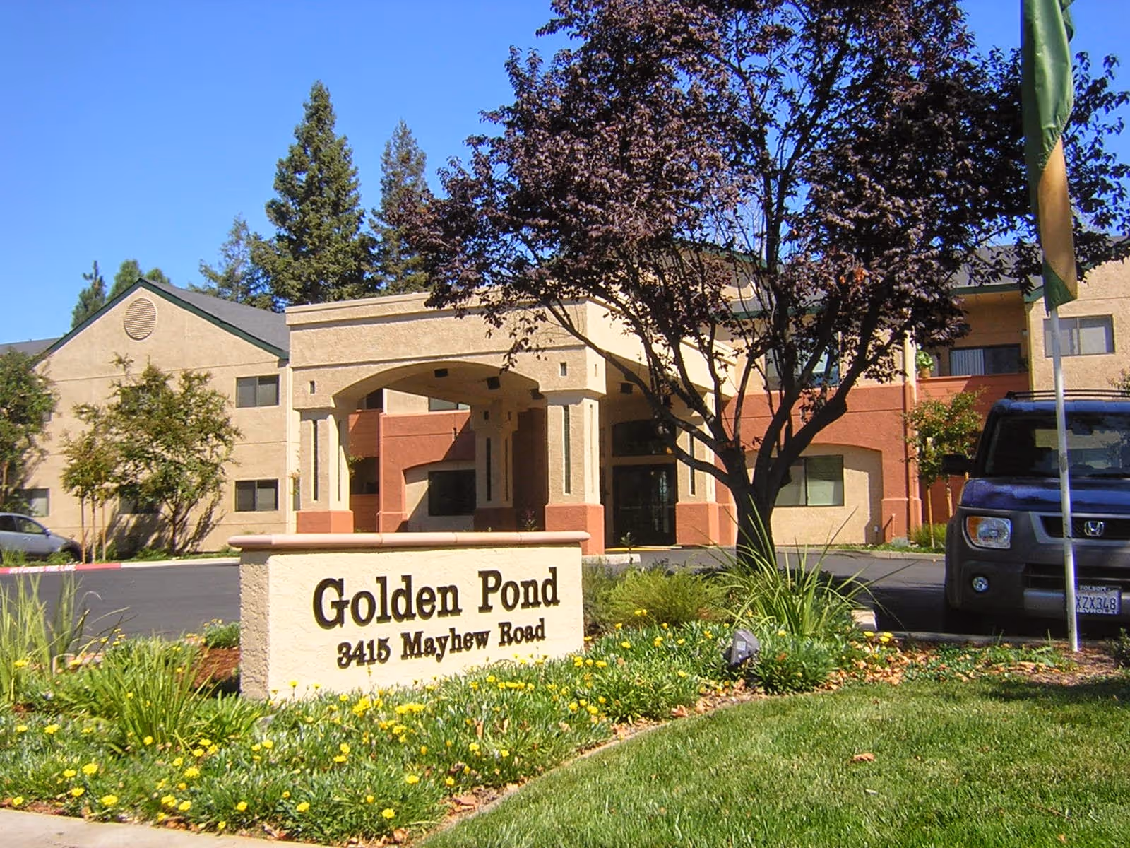 Exterior view of Golden Pond Retirement Community building with a sign in front displaying the name and address 3415 Mayhew Road. The building is two stories with beige and reddish walls, surrounded by trees, plants, and a parked car.