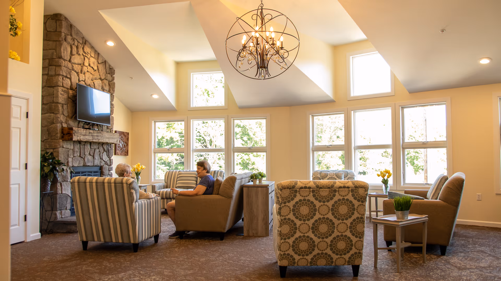 A bright and spacious living room with large windows letting in natural light. The room features a stone fireplace with a mounted TV above it, several comfortable armchairs and sofas arranged in a conversational grouping, small side tables with plants and flowers, and a decorative chandelier hanging from the ceiling. Two people are seated and engaged in conversation.