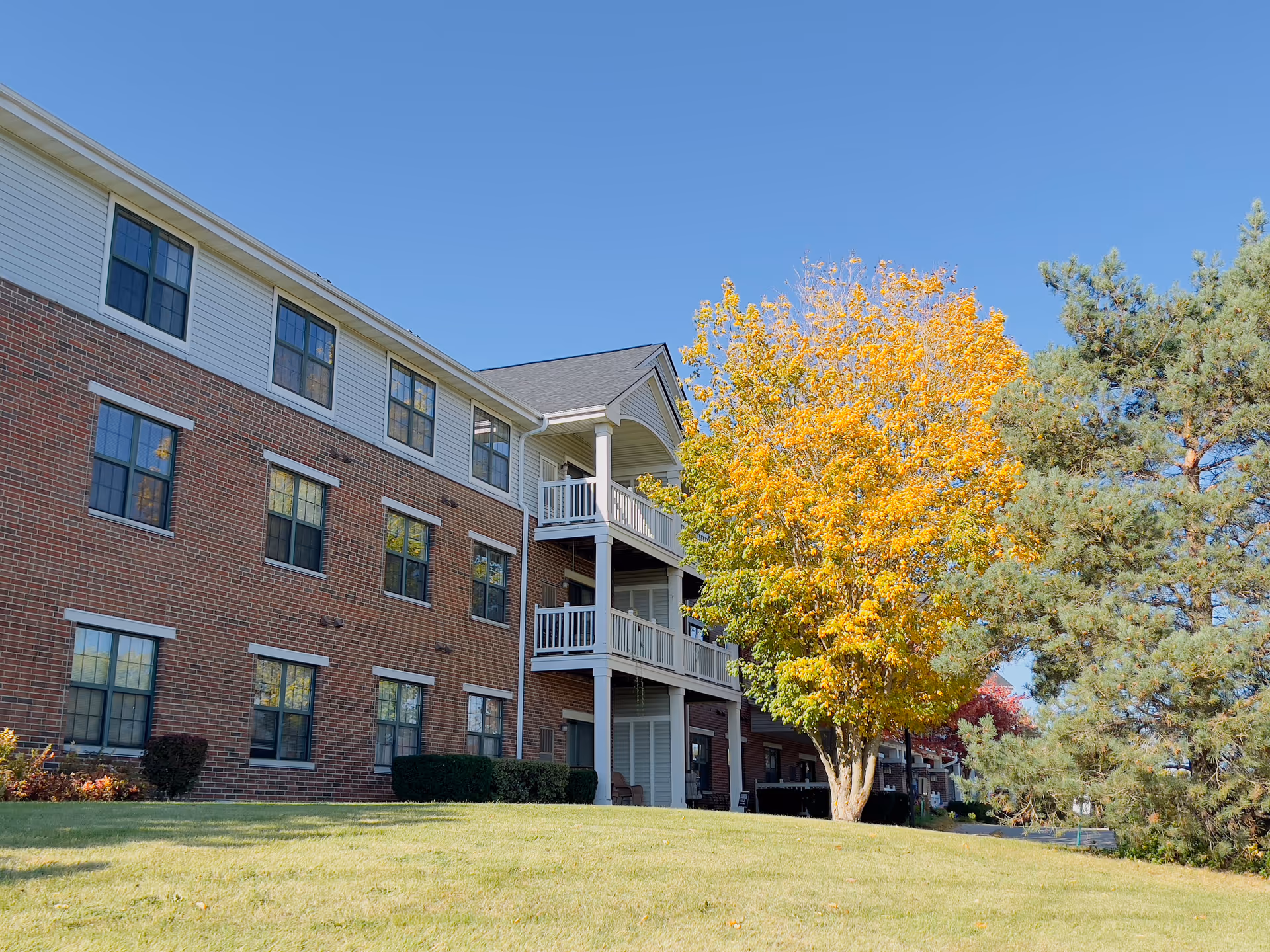 Three-story brick apartment building with balconies, a grassy lawn, and a tree with yellow autumn leaves under a clear blue sky.