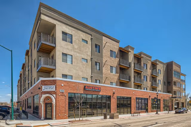 Exterior view of a multi-story residential building with a brick and beige facade under a clear blue sky. The ground floor features large windows and a sign for Austin's American Grill. The building has balconies on the upper floors and is located on a street corner with a sidewalk and street lamps.