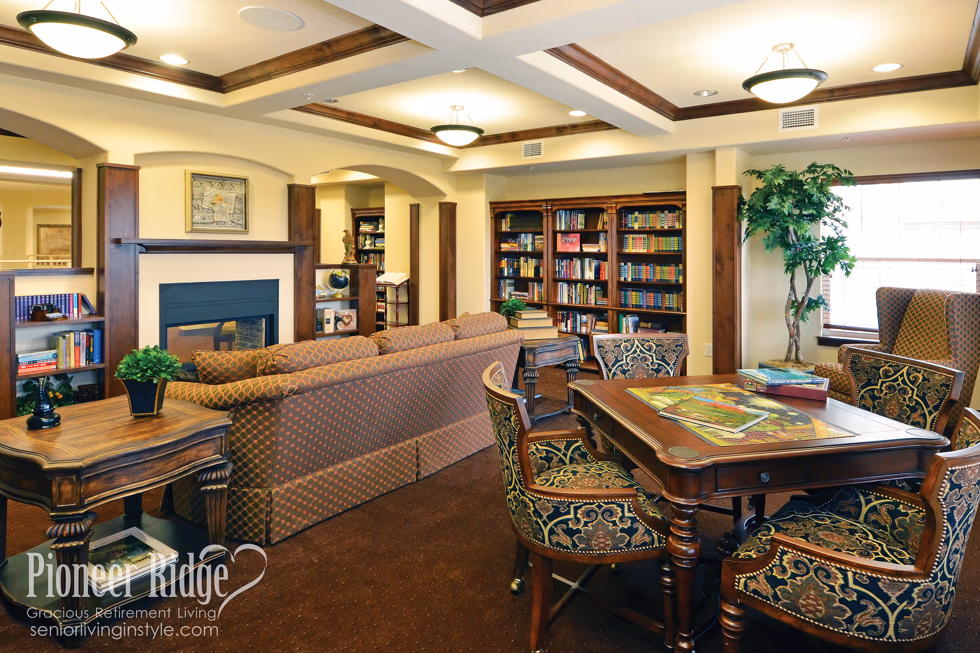 Cozy senior living common room with bookshelves, a fireplace, patterned sofas and a wooden game table by a window.