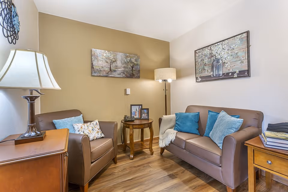 A cozy living room with a brown leather armchair and sofa, both adorned with blue and patterned throw pillows. There is a wooden side table with a lamp and another round wooden table with a telephone and framed photos. The walls are decorated with two paintings, one of trees and the other of flowers in a vase. The floor is wooden, and the room is softly lit by a floor lamp and a table lamp.