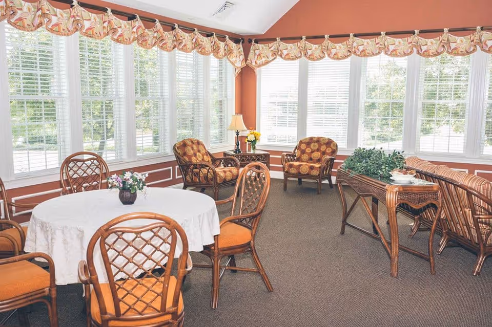 Bright sunroom-style common area with a round table, wicker chairs and upholstered armchairs in front of large windows.