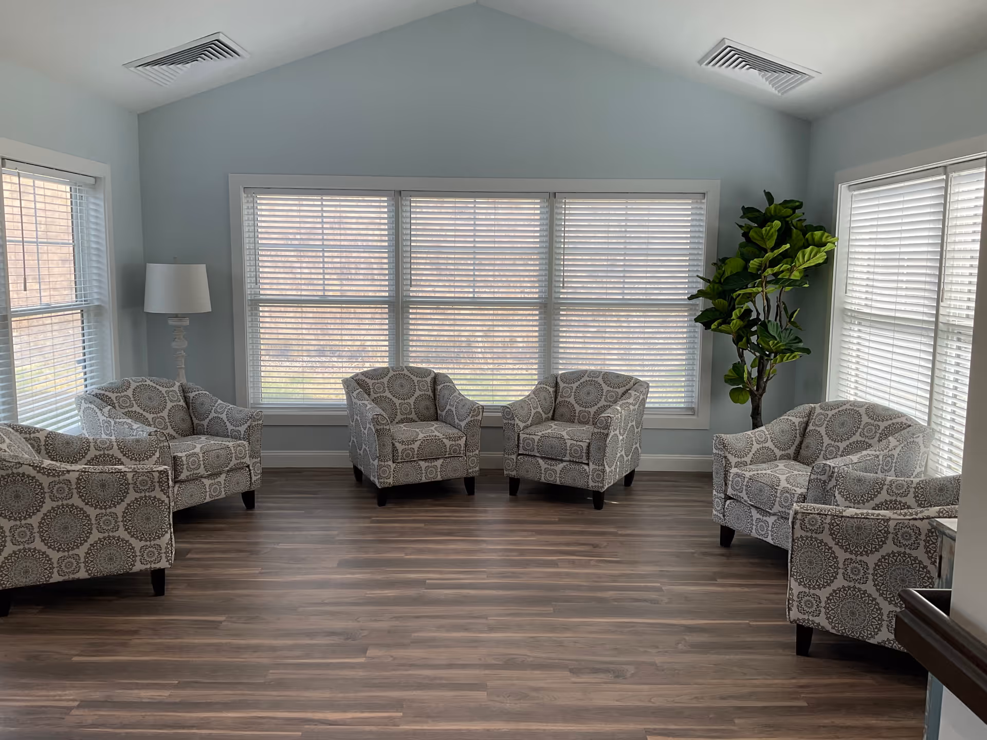 Bright seating room with several patterned armchairs arranged around large windows, wood flooring, a floor lamp, and a potted plant.