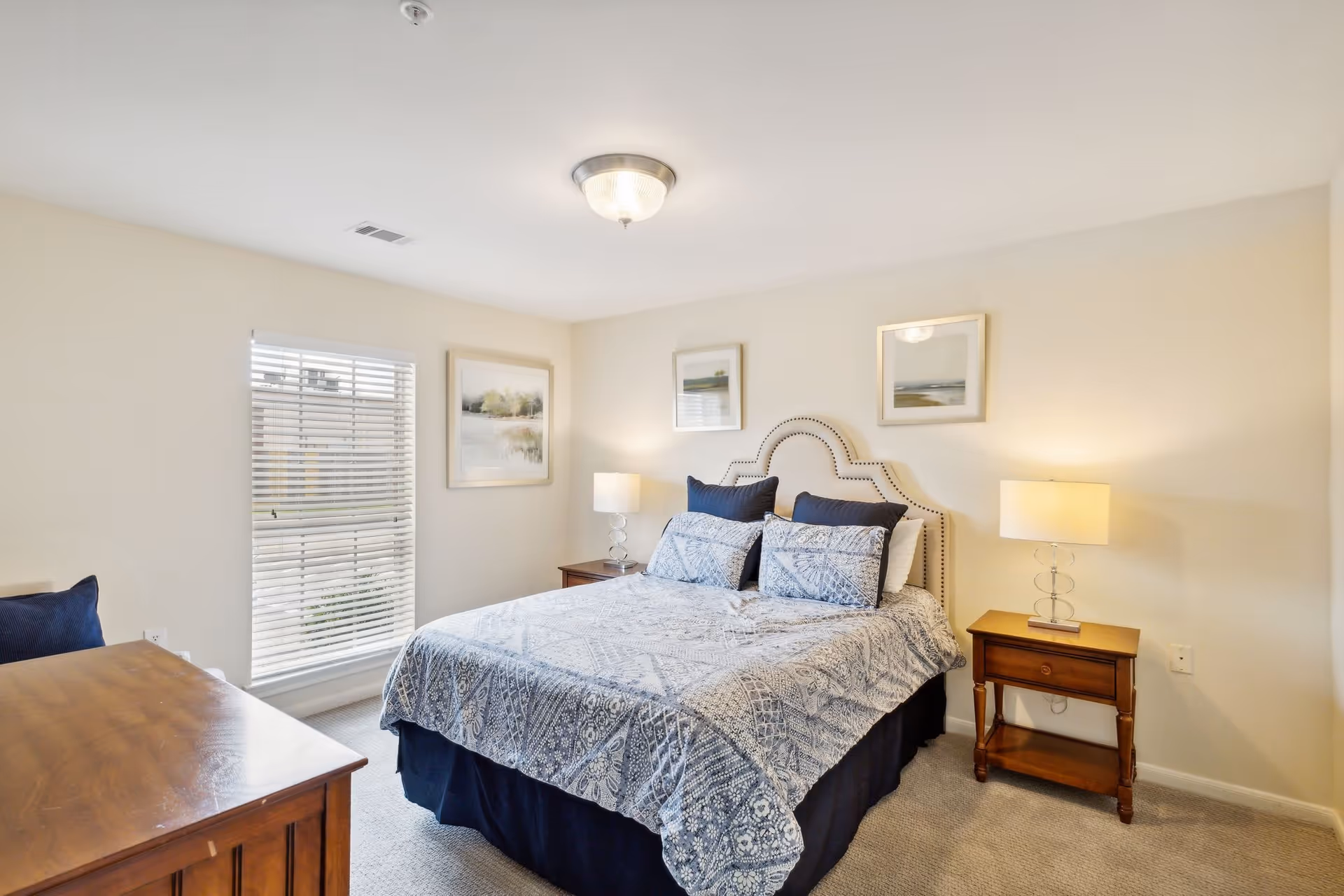 A bedroom with a neatly made bed featuring a patterned blue and white bedspread and matching pillows. The bed has a decorative headboard with nailhead trim. There are two wooden nightstands on either side of the bed, each with a lamp. The room has beige walls, a window with white blinds, and three framed landscape paintings on the walls.