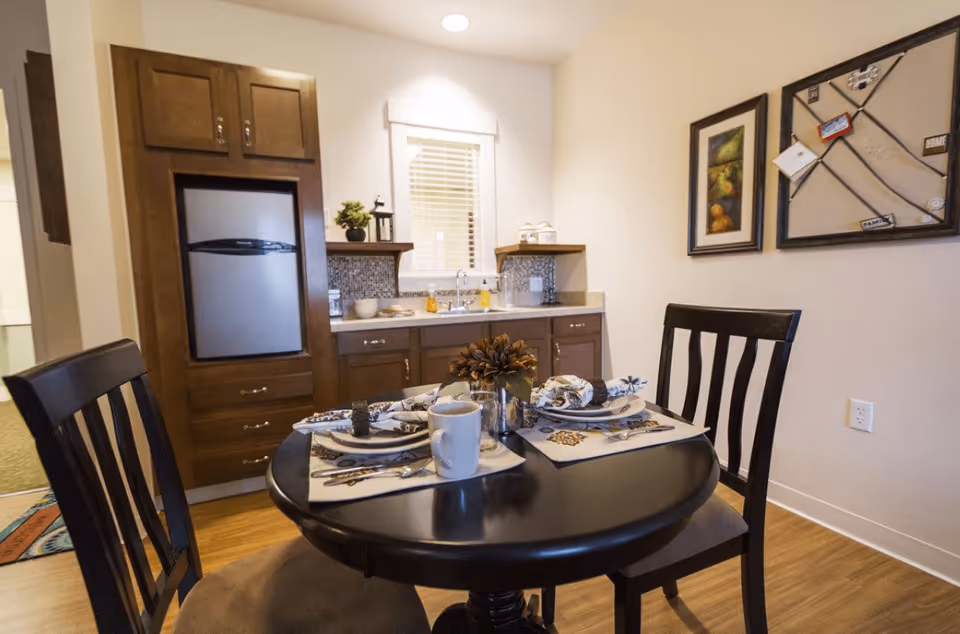 A small dining area with a round black table set for two, including plates, napkins, utensils, and a white mug. Behind the table is a kitchenette with a small refrigerator, cabinets, a sink, and a window with blinds. The walls are decorated with two framed pictures and a bulletin board with notes and photos.