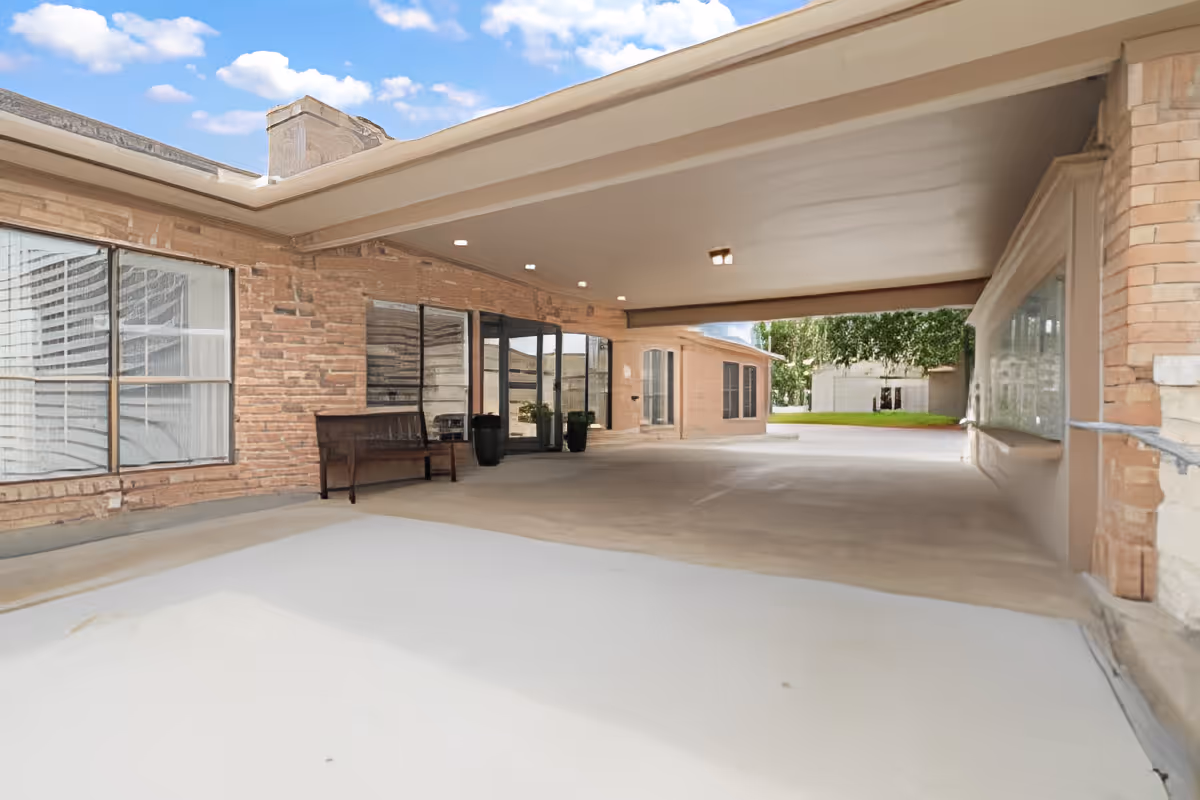 Covered outdoor entrance area of a building with brick walls, large windows, a bench, and potted plants. The area opens to a driveway and a grassy yard with trees in the background under a partly cloudy sky.