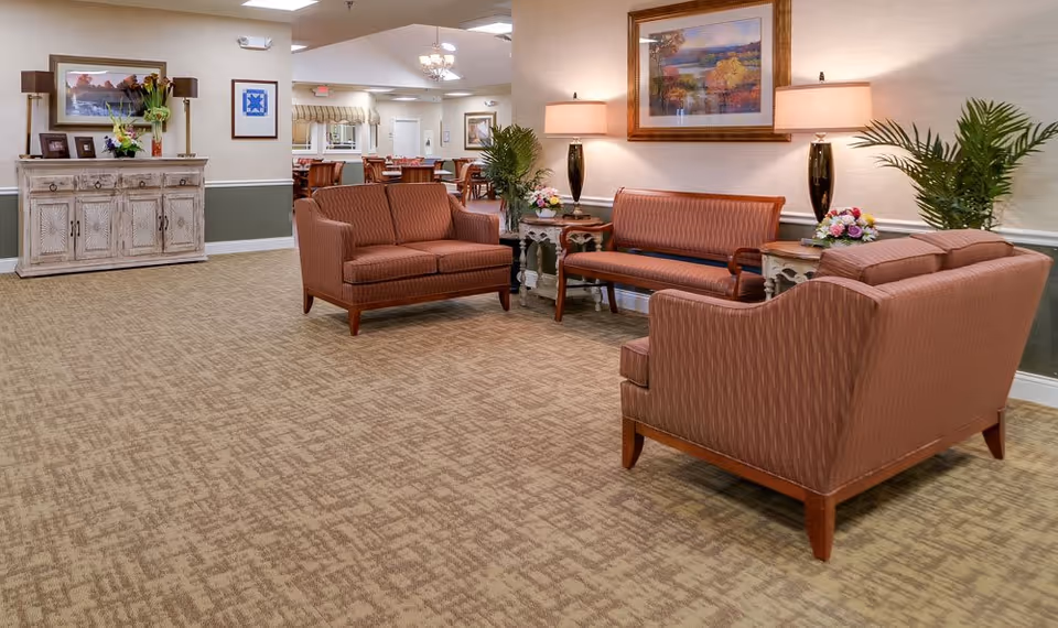 A cozy senior living common area with three upholstered sofas arranged around two small side tables with lamps and flower arrangements. The room features beige patterned carpet, light-colored walls with a green wainscot, framed artwork, and decorative plants. In the background, a dining area with tables and chairs is visible.