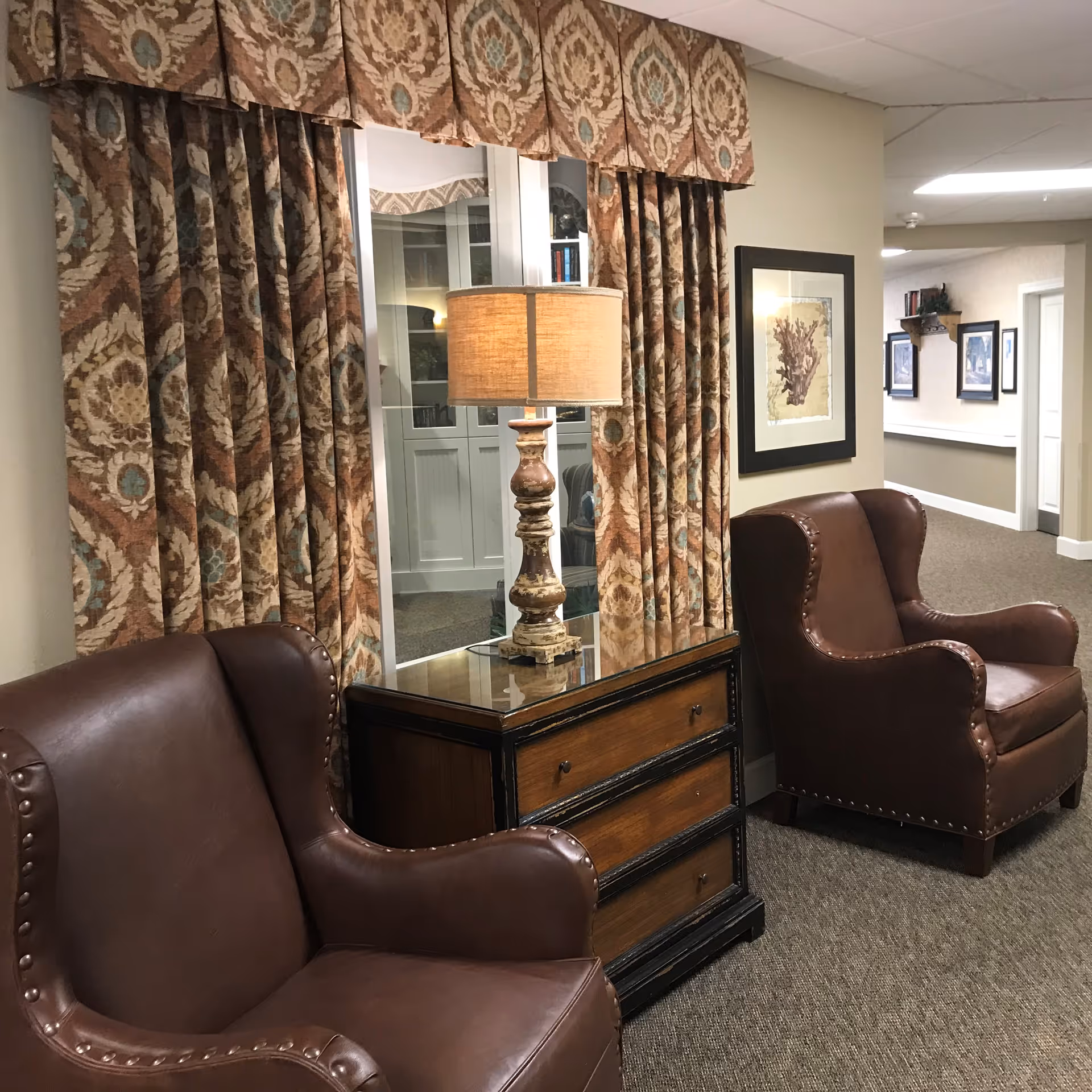 A cozy seating area in a hallway with two brown leather armchairs separated by a wooden side table with a decorative lamp. Behind the table is a window with patterned curtains. The hallway walls are adorned with framed artwork and the carpet is a neutral tone.