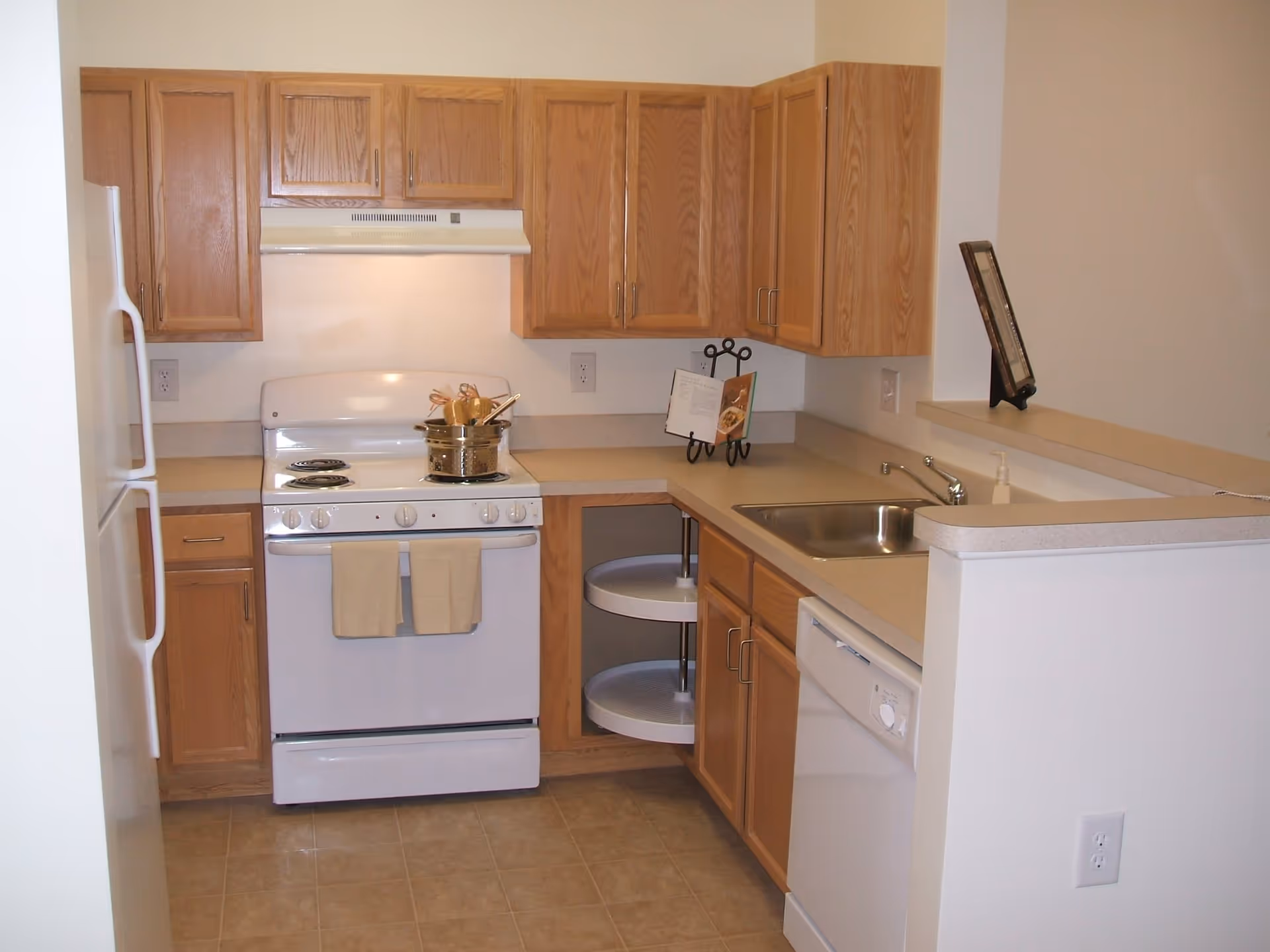 A compact kitchen with wooden cabinets, a white electric stove with two beige towels hanging on the handle, a white refrigerator, a stainless steel sink, a white dishwasher, and a corner cabinet with rotating shelves. There is a cookbook stand with an open cookbook on the countertop.