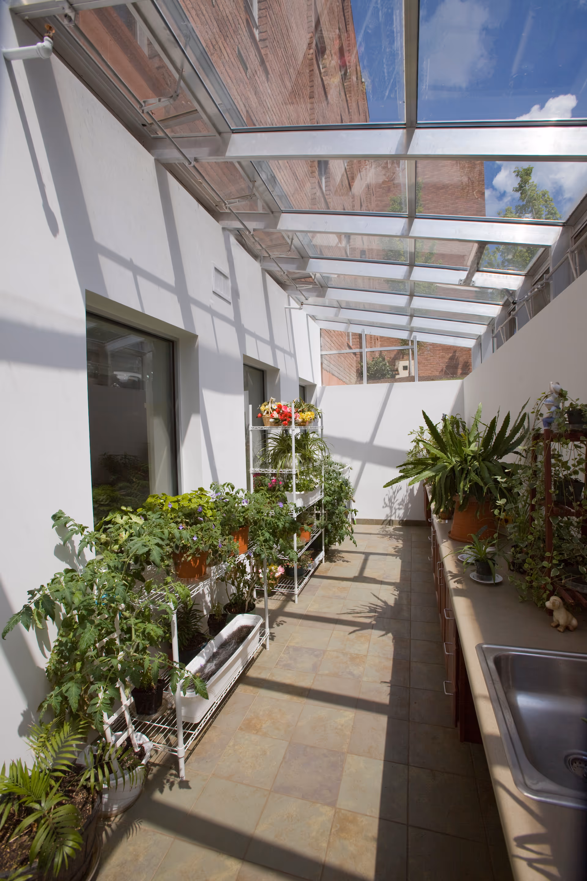 A bright indoor garden room with a glass ceiling allowing sunlight to stream in. The room has white walls and tiled floor, with various potted plants and flowers arranged on metal shelves and a countertop with a sink on the right side.