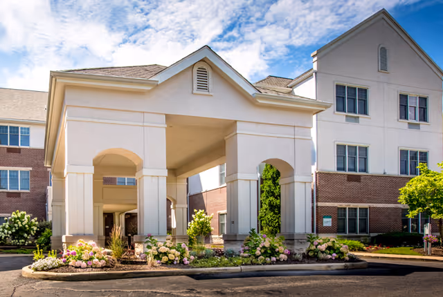 Exterior view of a senior living facility named Forest Hills Place featuring a covered entrance with columns, landscaped flower beds with blooming hydrangeas, and a multi-story building with brick and light-colored siding under a partly cloudy sky.