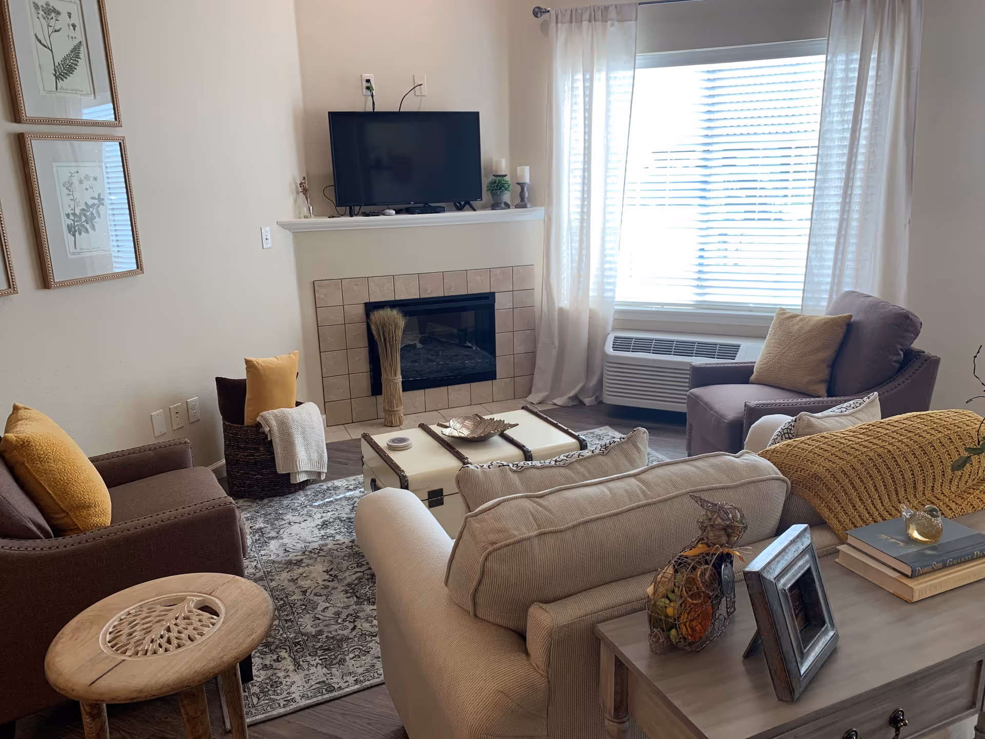 A cozy living room with a beige sofa and two brown armchairs, each with yellow cushions. A small wooden side table is next to one armchair. In the center, there is a white trunk-style coffee table on a patterned rug. A fireplace with a tiled surround is topped by a flat-screen TV. A large window with white curtains and blinds lets in natural light. Decorative items including framed botanical prints, a basket with pillows and a blanket, and a small table with books and a decorative bird sculpture complete the room.