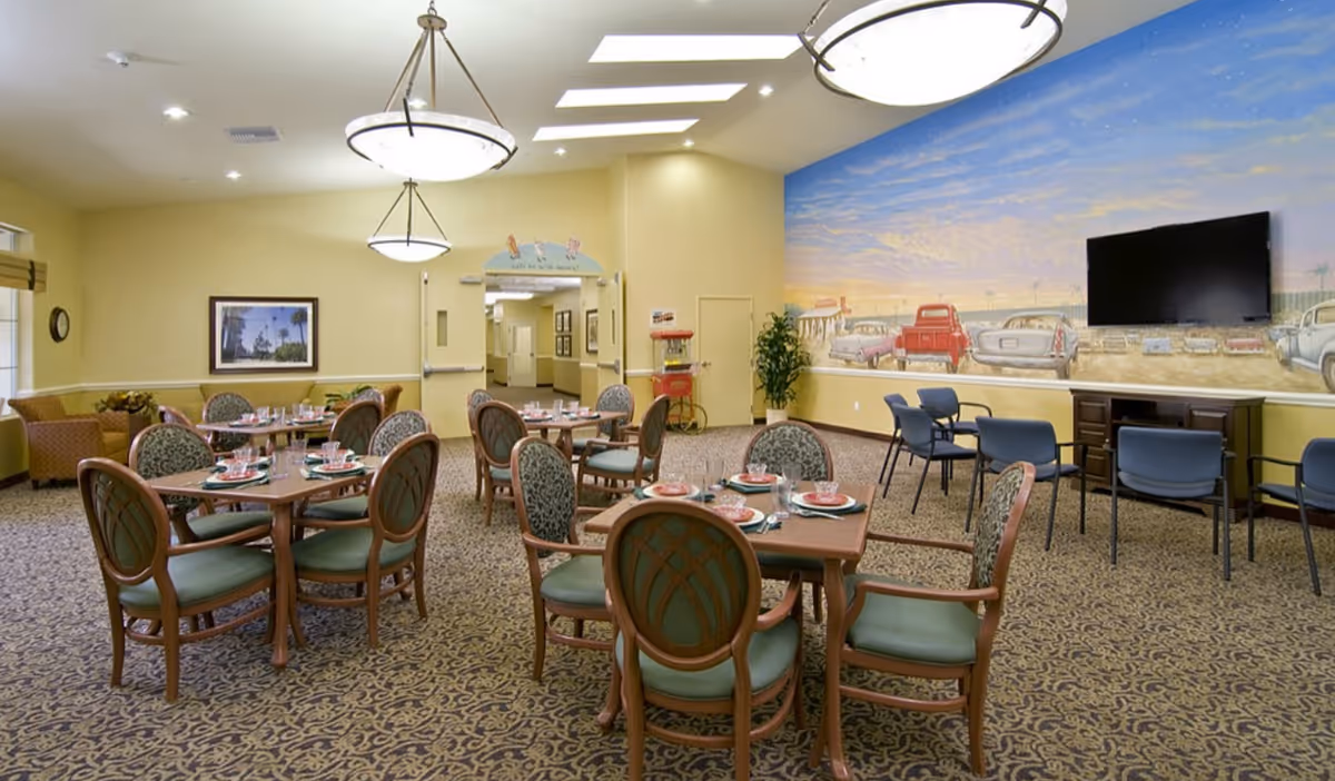 A dining room in a senior living facility with several wooden tables and cushioned chairs arranged neatly. Each table is set with plates, glasses, and utensils. The room has patterned carpet flooring, beige walls, and two large ceiling lights. A mural depicting vintage cars and a sunset sky decorates one wall, which also has a mounted flat-screen TV. There are additional chairs lined up against the wall beneath the TV and a popcorn machine near the entrance.