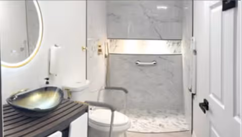 Modern bathroom with a round illuminated mirror, a stylish vessel sink on a wooden countertop, a white toilet, and a walk-in shower with marble walls and a grab bar.