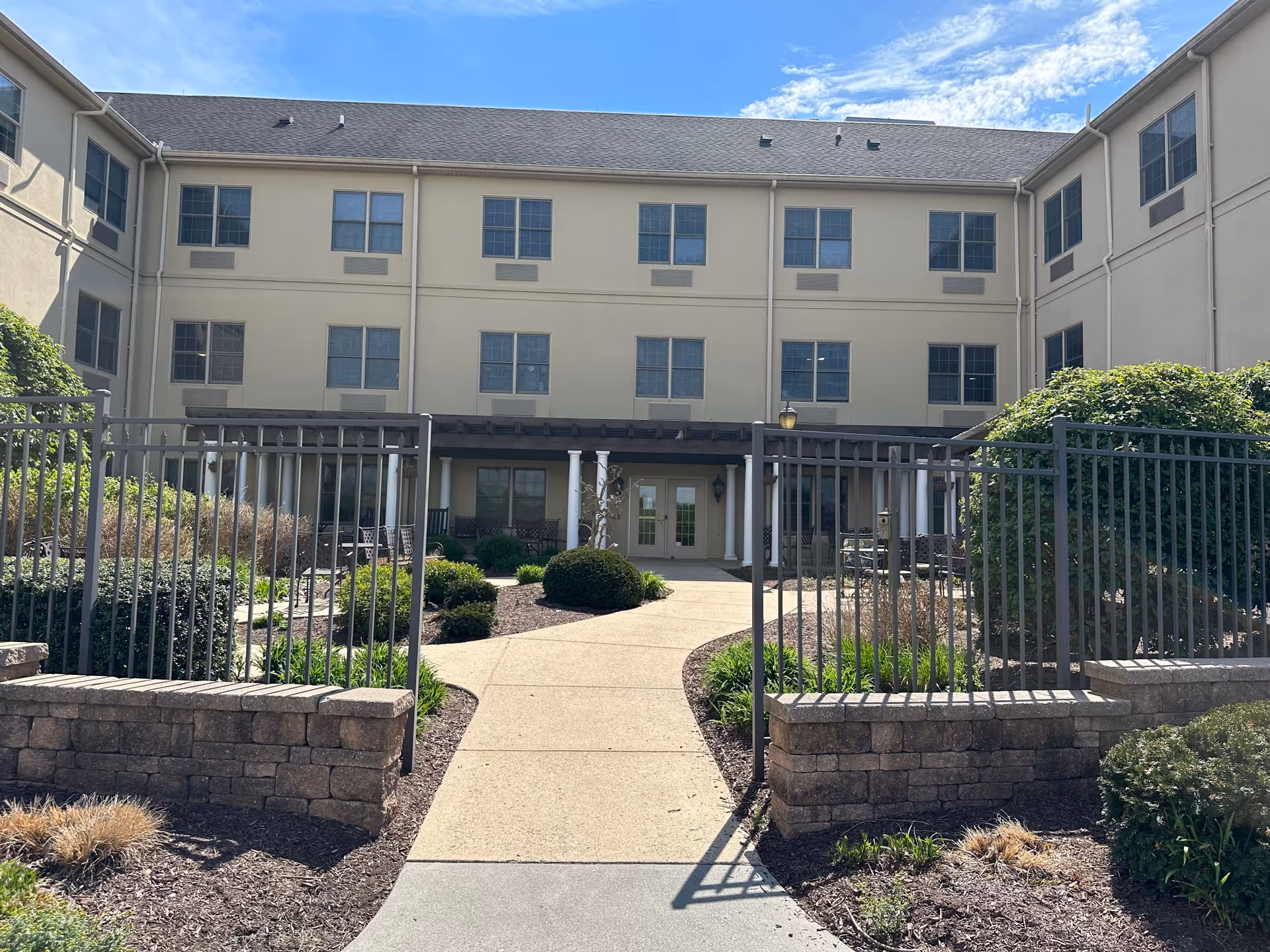Outdoor courtyard area of a multi-story retirement community building with a paved walkway leading through a garden with shrubs and plants, surrounded by a metal fence and stone retaining walls under a blue sky with some clouds.