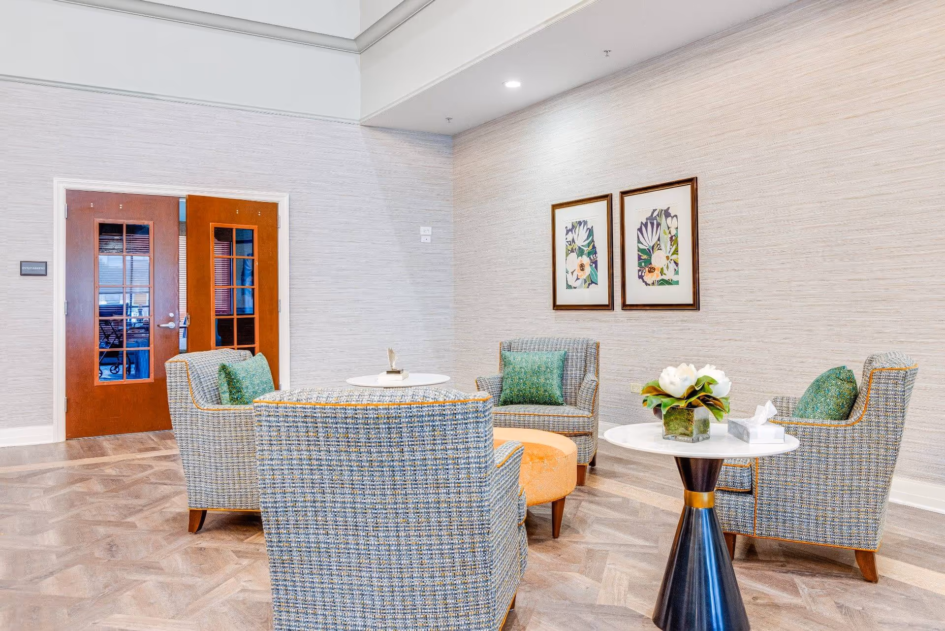 Bright lounge with four upholstered armchairs around small round tables, a flower arrangement, double wooden doors, and framed botanical prints on the wall.