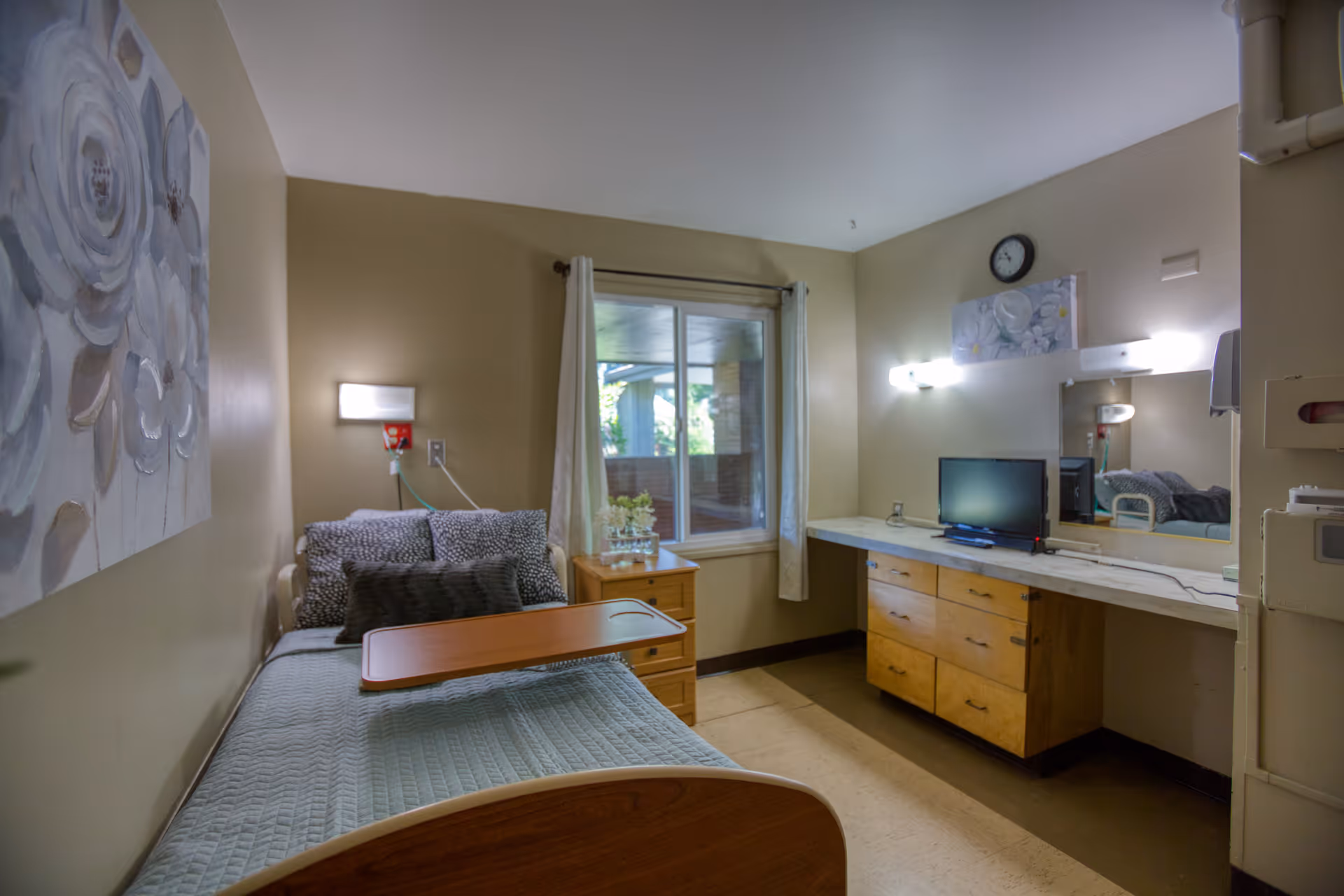 A small, neatly arranged bedroom in a senior living facility with a single bed featuring a light blue quilt and multiple pillows. A wooden overbed table is positioned on the bed. Next to the bed is a wooden nightstand with a small plant and a window with white curtains allowing natural light. Across from the bed is a long wooden dresser with a small flat-screen TV, a large mirror, and wall-mounted lights. The walls are painted beige and decorated with floral artwork.