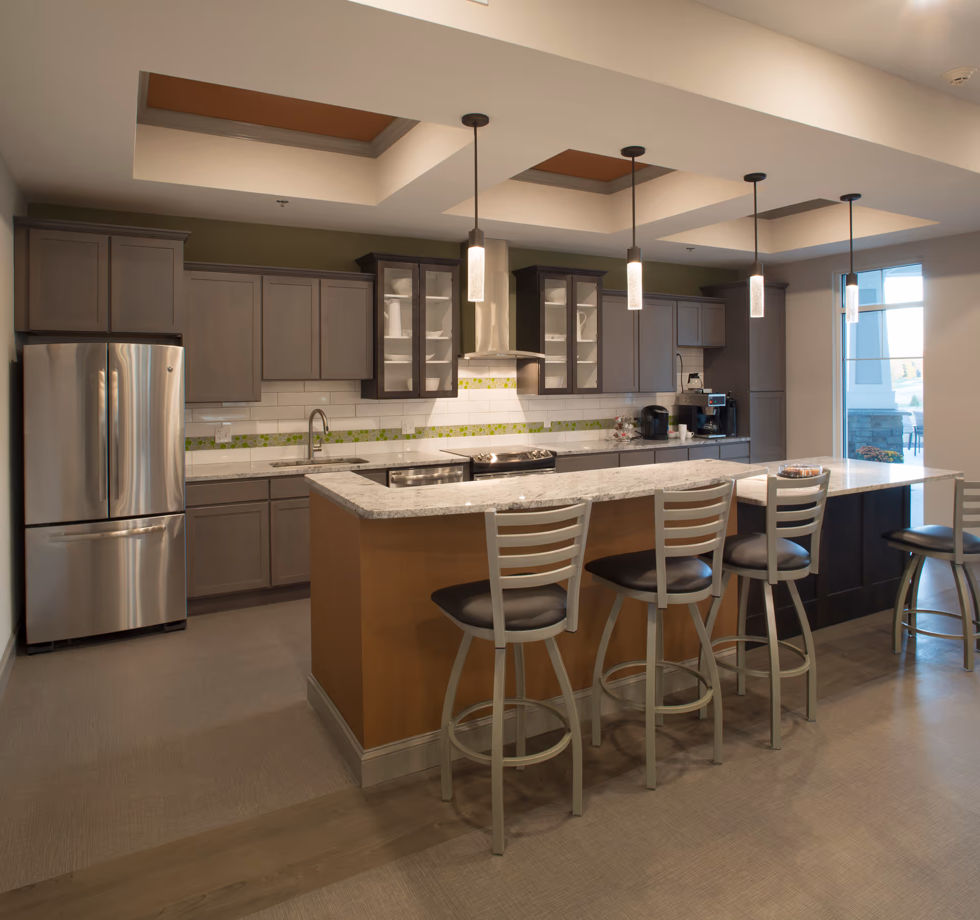 Modern kitchen area with a large island countertop featuring four bar stools. The kitchen has gray cabinets, stainless steel refrigerator, stove with a hood, and pendant lights hanging from the ceiling. There is a coffee machine on the counter and a window letting in natural light.