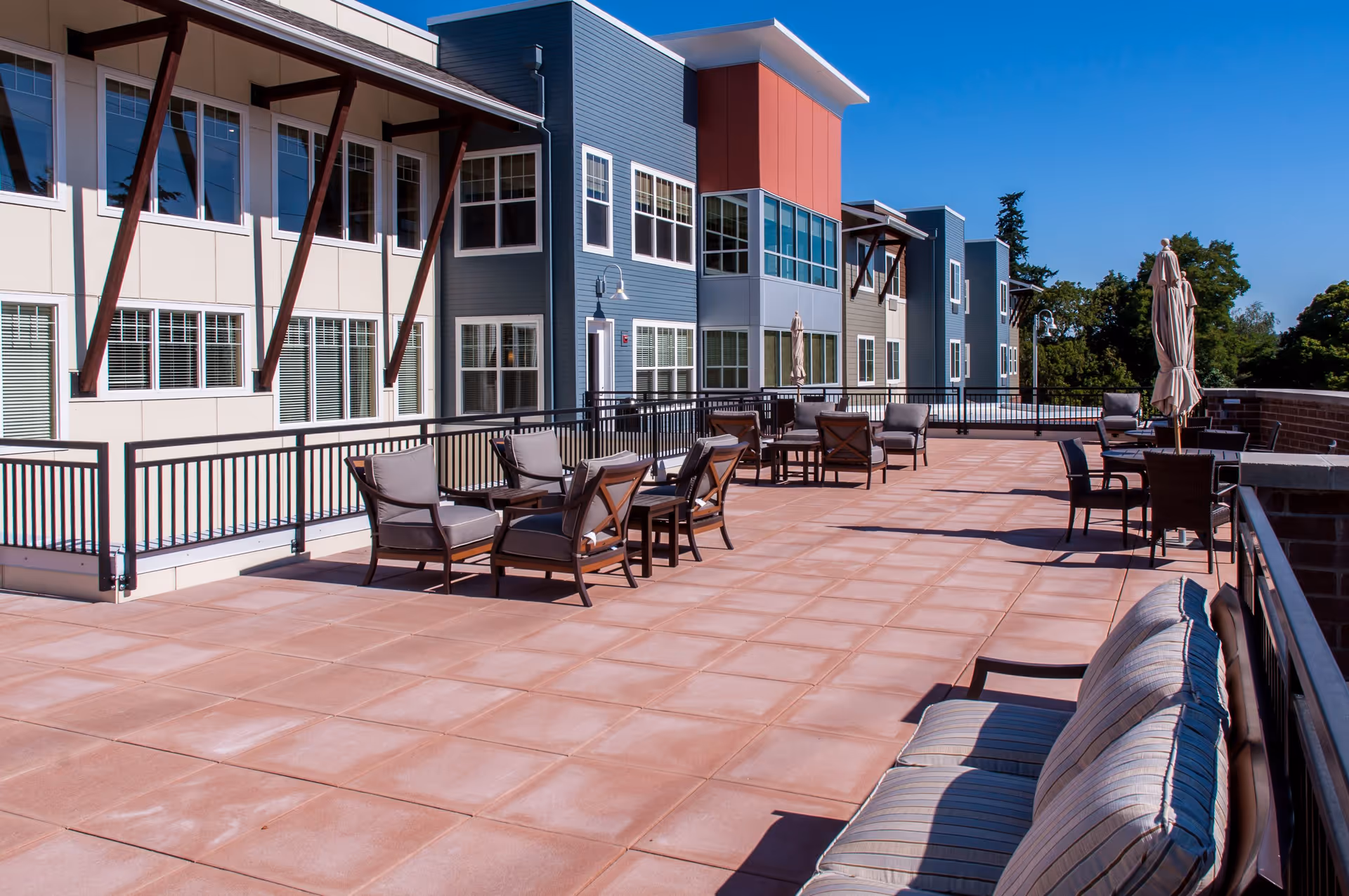 Outdoor patio area at Marquis Tualatin Assisted Living with multiple seating arrangements including cushioned chairs and tables, surrounded by a railing and adjacent to a multi-story building with large windows under a clear blue sky.