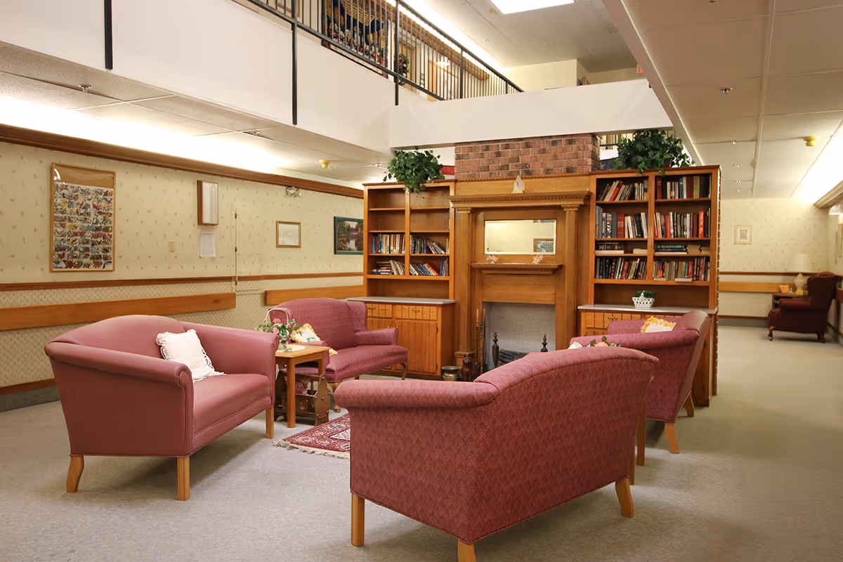 A cozy senior living facility lounge with pink upholstered sofas arranged around a central fireplace and bookcases.