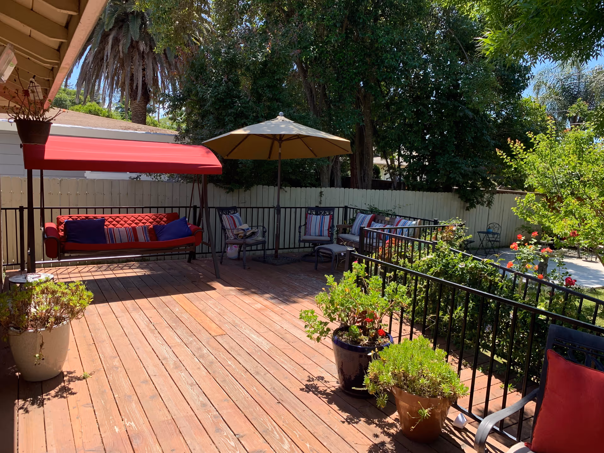Sunlit wooden deck with a red-canopy swing, outdoor umbrella and seating surrounded by potted plants and a fenced yard.