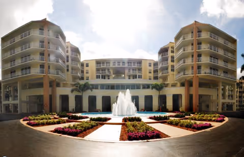 Wide view of a multi-story senior living facility named Park Summit with a central water fountain surrounded by landscaped flower beds and palm trees under a partly cloudy sky.