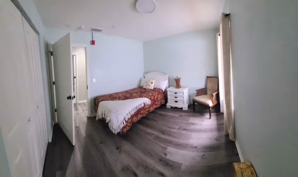 A small bedroom with a single bed covered in a red floral bedspread and a white knitted throw. Next to the bed is a white nightstand with a decorative item on top. There is a beige upholstered chair near a window with light-colored curtains. The room has light blue walls and dark wood flooring. A door is open, leading to another room or hallway.