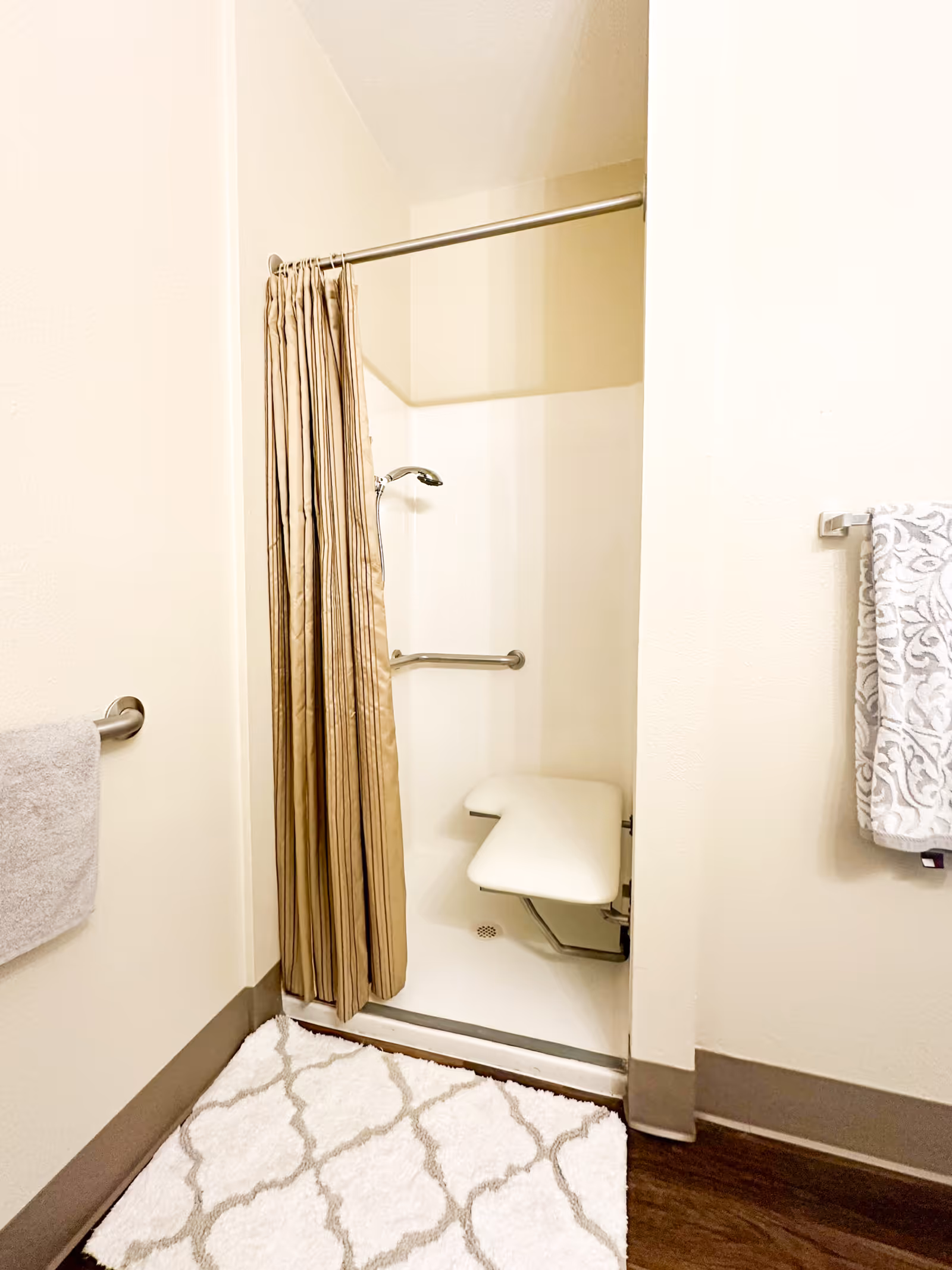 Accessible shower stall with a beige curtain, fold-down seat, grab bar, and towels in a bathroom.