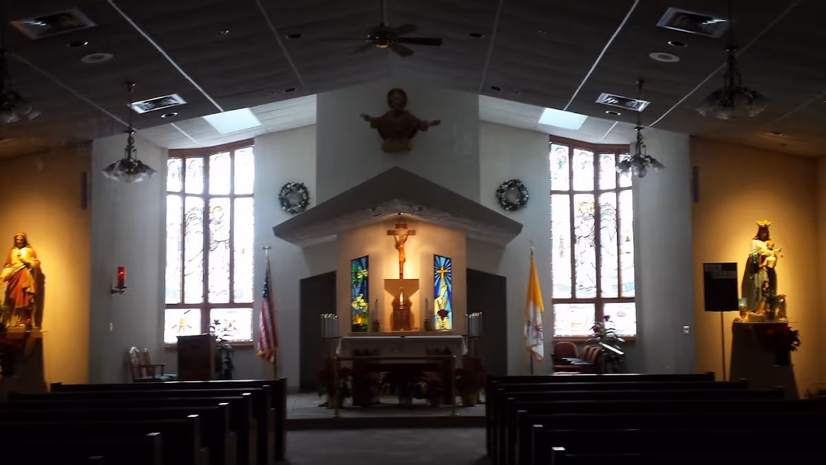 Interior view of a chapel with wooden pews facing an altar. The altar features a crucifix, stained glass windows on either side, and religious statues on both left and right walls. The ceiling has recessed lighting and ceiling fans, and there are American and Vatican flags near the altar.