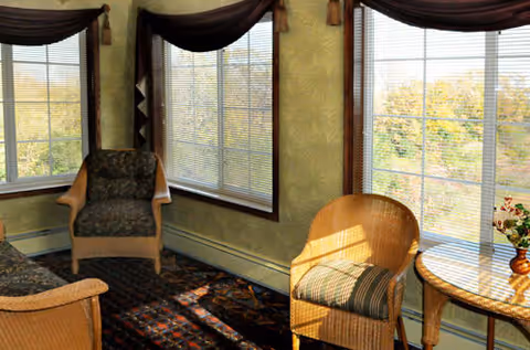 A sunlit sitting area with large windows featuring horizontal blinds. The room contains a cushioned armchair, a wicker chair with a striped cushion, a small round table with a vase of flowers, and a patterned rug on the floor. The walls have a light greenish wallpaper with a subtle pattern, and dark curtains hang above the windows.