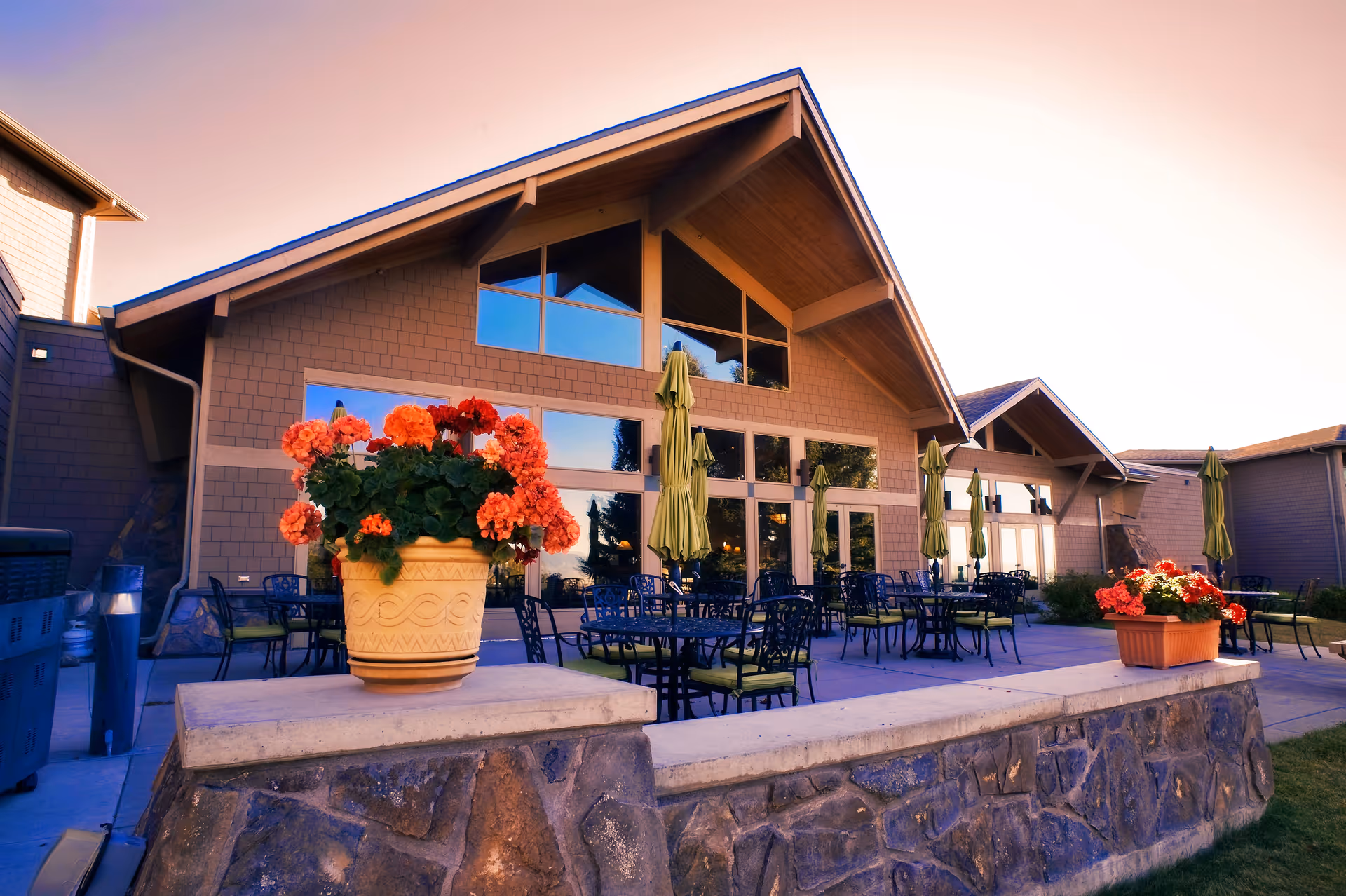 Outdoor patio area of Bozeman Health Hillcrest Senior Living, Aspen Pointe, featuring a stone wall with potted red and orange flowers, multiple tables and chairs with green cushions, and closed green umbrellas in front of a building with large windows and a peaked roof.