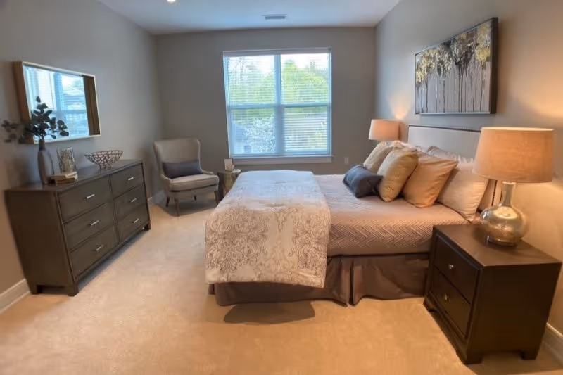 A neatly arranged bedroom with a large bed covered in a patterned comforter and multiple pillows. There are two bedside tables with lamps on either side of the bed. A dresser with decorative items and a mirror is positioned against the left wall. A comfortable armchair with a cushion and a small side table are placed near a window with blinds, allowing natural light to brighten the room.