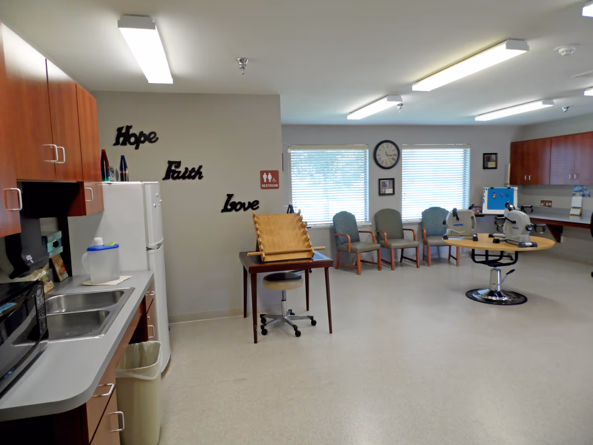 A bright room with a kitchenette on the left featuring a sink, microwave, and refrigerator. On the wall above the refrigerator are decorative words 'Hope', 'Faith', and 'Love'. In the center of the room is a small table with a wooden pegboard game and a stool. Against the far wall are several chairs arranged in a row beneath two windows with blinds. A round table with two exercise machines is positioned near the chairs. The room has overhead fluorescent lighting and a clock on the wall between the windows.