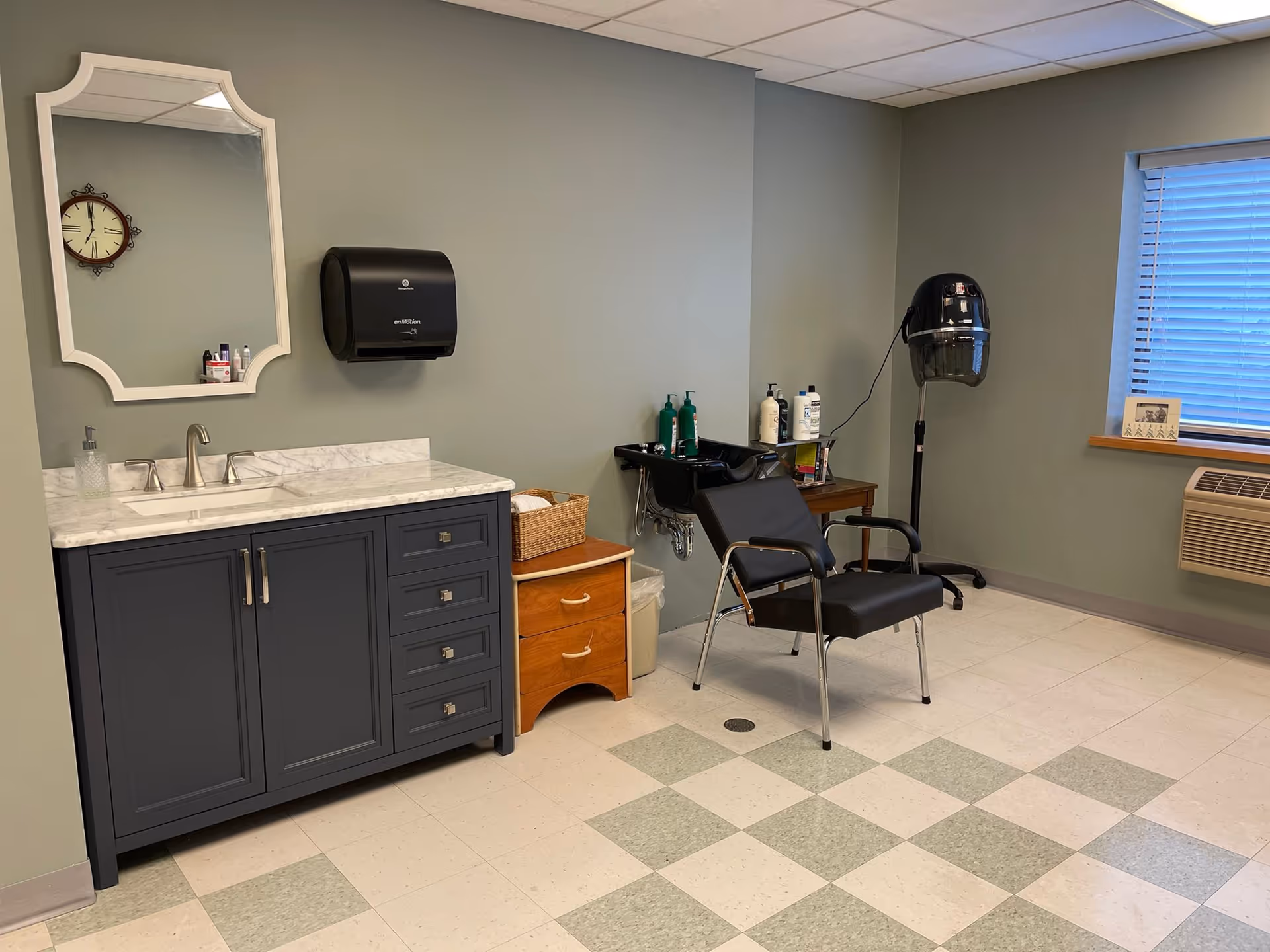 Small salon-style interior with a marble-top vanity and mirror on the left, a shampoo sink and salon chair with a hood dryer nearby, and a window air conditioner.