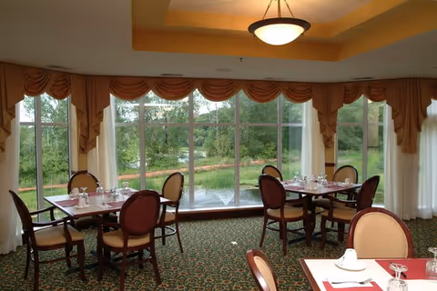 A dining room with several tables and chairs arranged neatly. Each table is set with white napkins, glasses, and silverware. Large windows with draped curtains provide a view of a green outdoor area with trees and a water fountain.