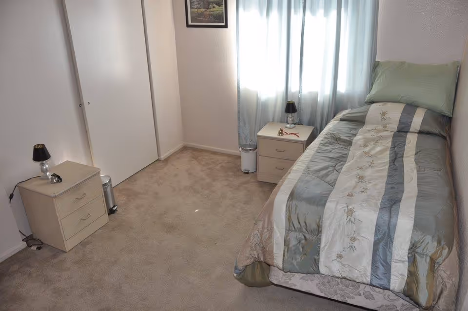 A small bedroom with a single bed covered in a green and beige striped comforter with floral patterns. There are two matching nightstands on either side of the bed, each with a small lamp. The room has beige carpet, white walls, a window with light blue curtains, and a closed white closet door.