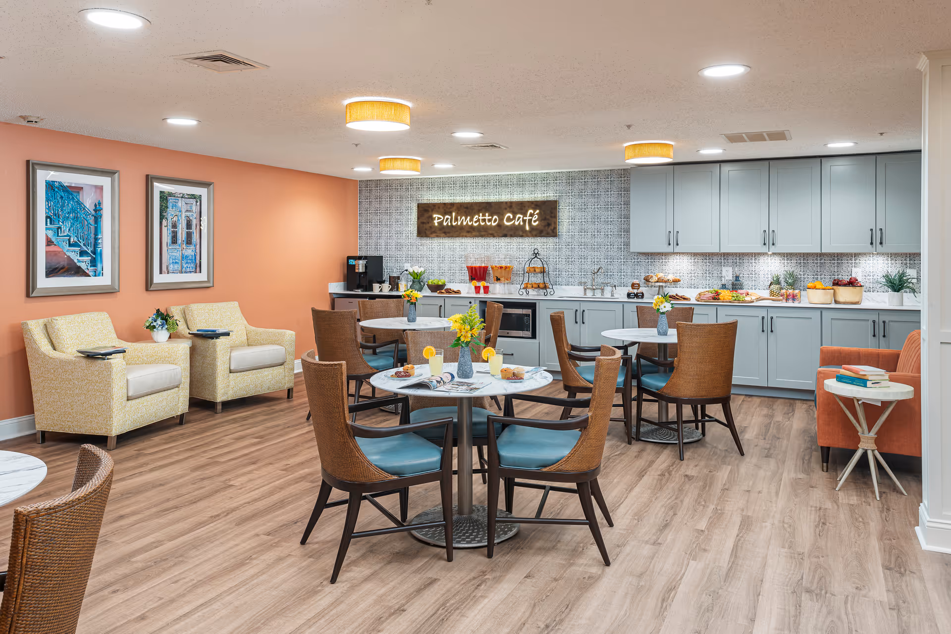 A cozy café area named Palmetto Café with round tables and chairs, a countertop with cabinets, a coffee machine, and various snacks and drinks. The room has warm lighting, framed artwork on a peach-colored wall, and comfortable seating including armchairs and a small sofa with a side table holding books.