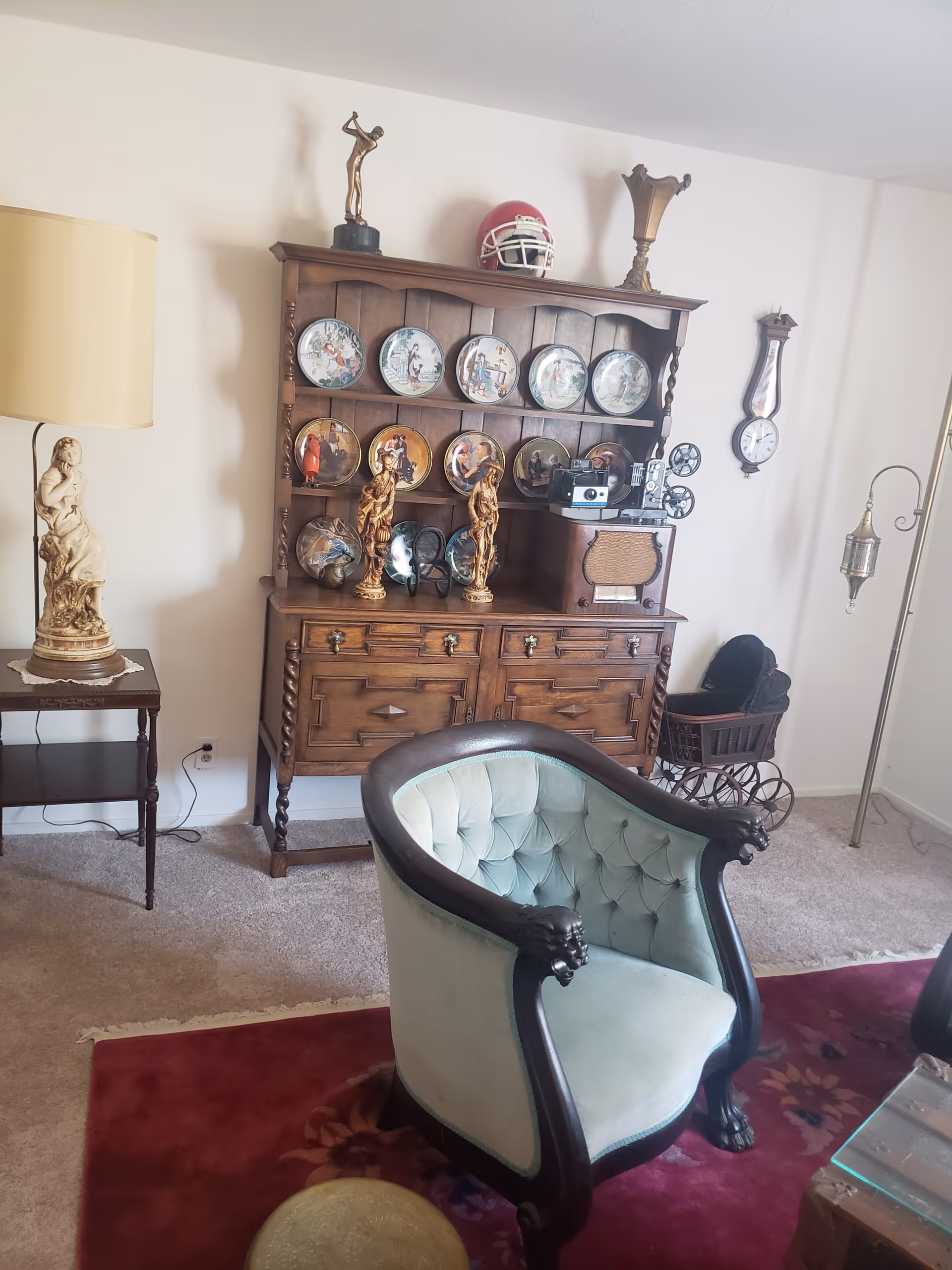 A cozy living room area featuring a vintage wooden cabinet displaying decorative plates and figurines, an old-fashioned radio, and a vintage camera. In front of the cabinet is a light blue upholstered armchair with dark wooden trim. To the left, there is a small side table with a lamp and a statue. The room has a carpeted floor with a red area rug and a wall clock hanging on the right wall.