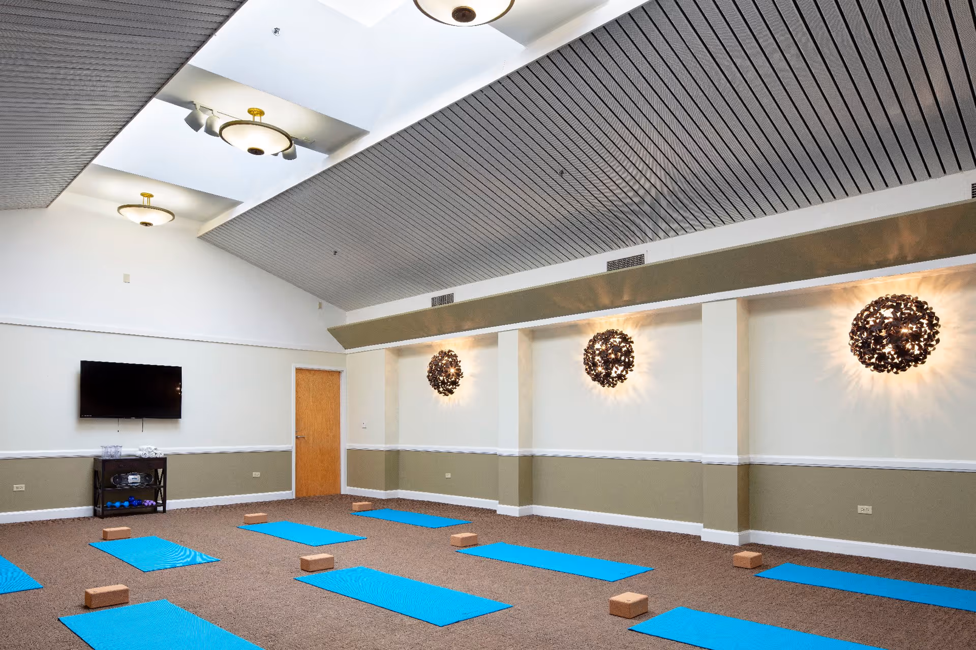 A spacious room with a high ceiling and modern light fixtures, set up for a yoga or exercise class with blue mats and wooden blocks evenly spaced on a carpeted floor. A flat-screen TV is mounted on one wall above a small table with water bottles and equipment.