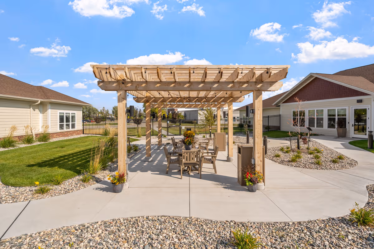 Outdoor patio area at Fargo Elim Health Care Center featuring a wooden pergola with string lights, several chairs and tables underneath, surrounded by landscaped pathways, grass, and buildings under a blue sky with scattered clouds.